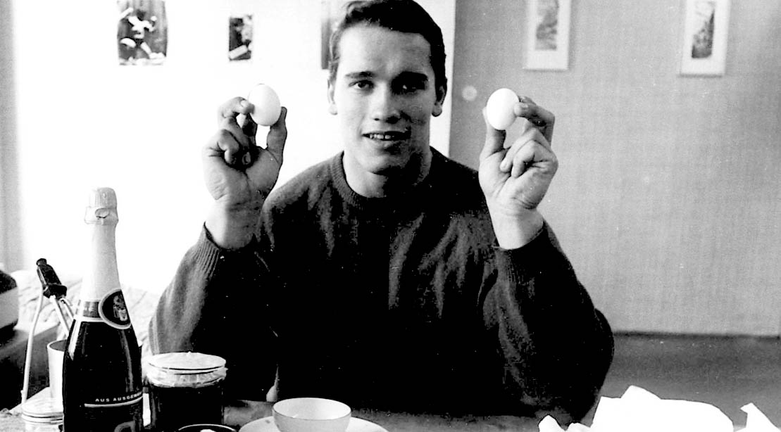 A young Bodybuilding Legend Arnold Schwarzenegger holding two hard boiled eggs for his diet for a bodybuilding physique