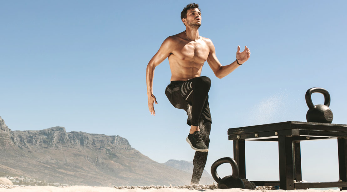 Physically fit man doing exercises on the beach with a kettlebell and high knee exercise