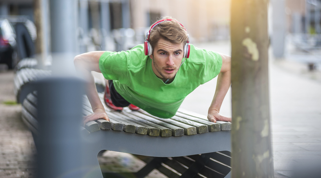 Young Sporty Man With Headphones Doing Pushups On Bench