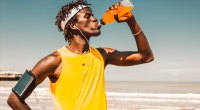 Fit runner drinking orange sports drink with electrolytes while running on the beach for hydration