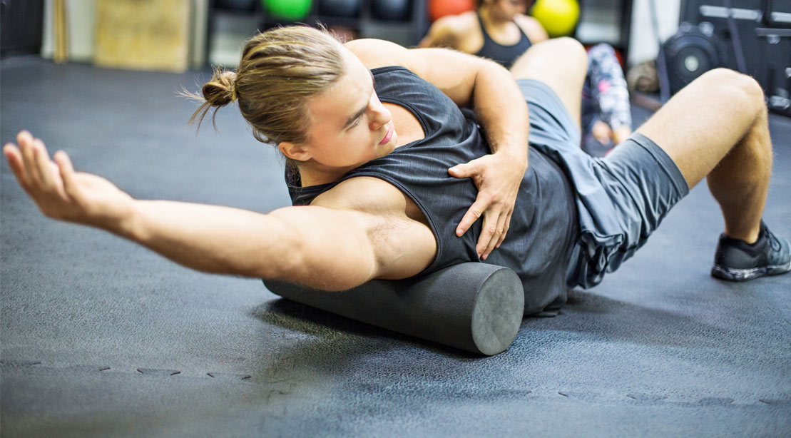 Male bodybuilder with a man bun performing foam rolling exercises on his arms with a foam roller