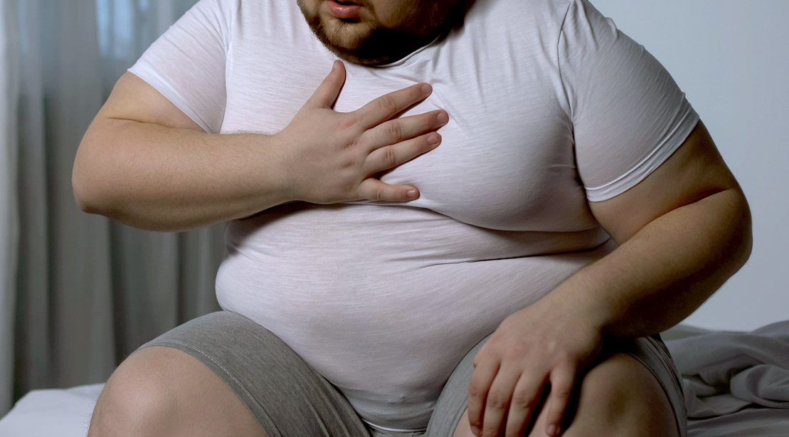 Overweight man with gynecomastia or man boobs sitting in his bed holding his chest