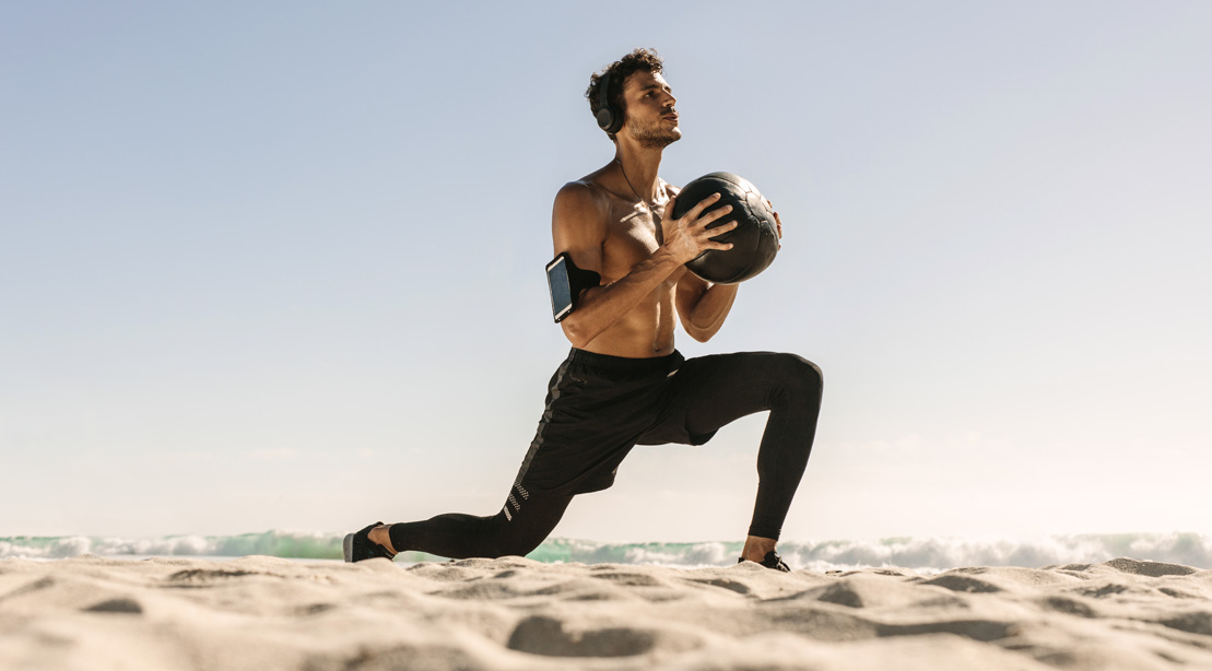 Fit man doing lunges with a medicine ball on the beach for his med ball training exercises