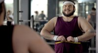Happy and overweight man who lost weight from his fitness resolution measuring his stomach in the gym mirror