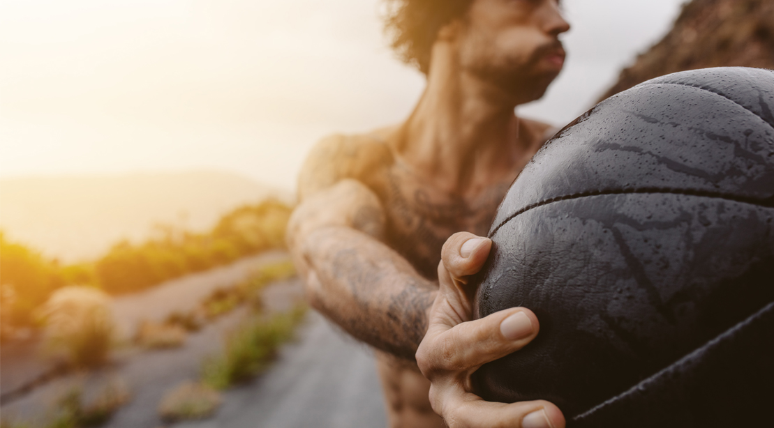 Fit muscular man performing lateral exercises using a medicine ball during sunrise on an empty road