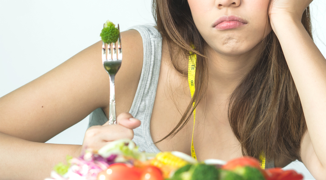 Unhappy girl eating a plate of vegetables with a fork.