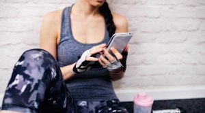 Female-In-Fitness-Clothing-Sitting-Against-Wall-On-Smart-Phone