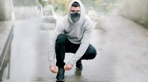 Man wearing a hoodie outdoors tying his sneakers while wearing a face mask during pandemic