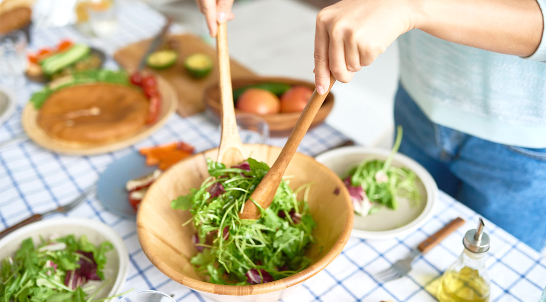 Woman serving herself a plate of mixed green salad from a wooden bowl.