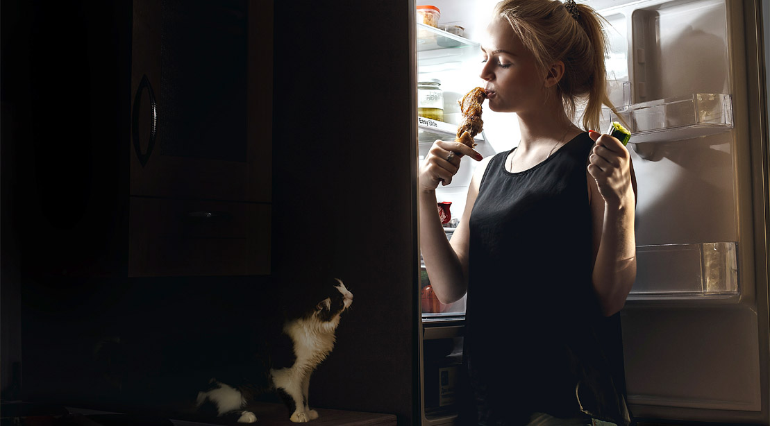 Girl eating chicken drumstick from the fridge while cat watches