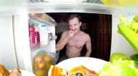 Mann isst bis spät in die Nacht kalte Pizza vor einem Kühlschrank, während sein alternder Stoffwechsel langsamer wird