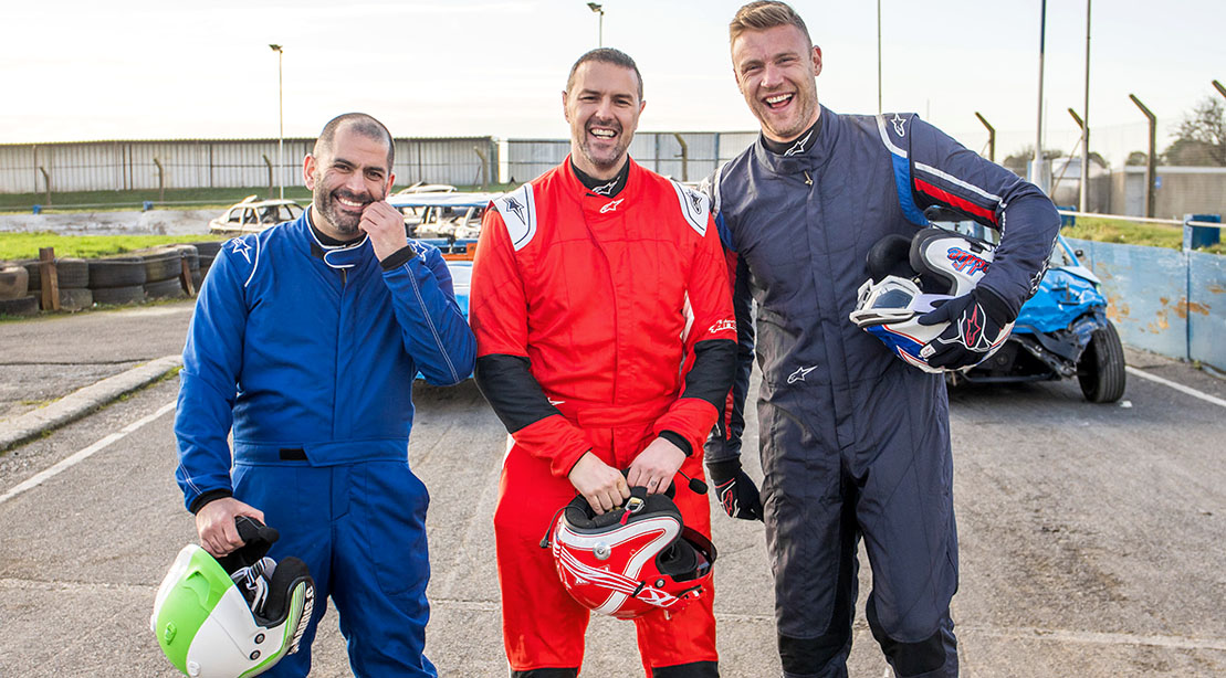 Top Gear hosts Chris Harris, Paddy McGuinnes, and Andrew Flintoff standing on a car race track in driving suits