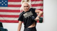 Blonde female model performing a dumbbell clean in front of the USA flag for Project Rock “For The Heroes” Collection