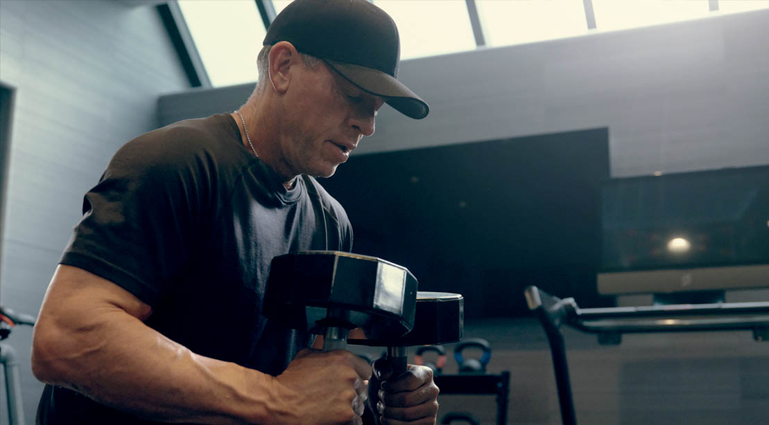Former NFL Cowboys quarterback hall of famer Troy Aikman working out in the gym doing a dumbbell exercise at the age of 55