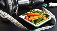 Bowl of steamed vegetables next to dumbbells and measuring tape