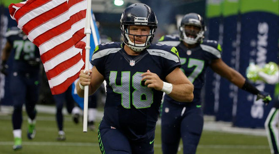 Nate Boyer wearing a seattle seahawks uniform and holding the American Flag