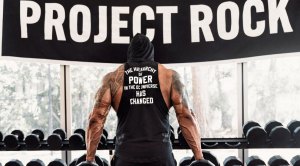 The Rock in front of the dumbbell rack with a Project Rock banner