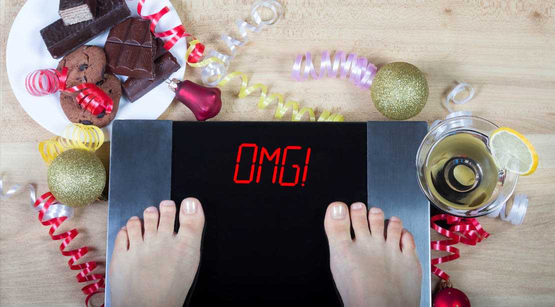 Woman on a digital scale displaying OMG with holiday food surrounding the scale for a fit christmas